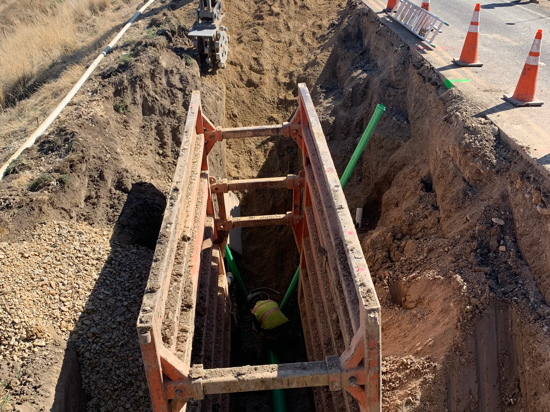 Trench with shoring for underground utility work. Green pipes are visible. Cones line the road.