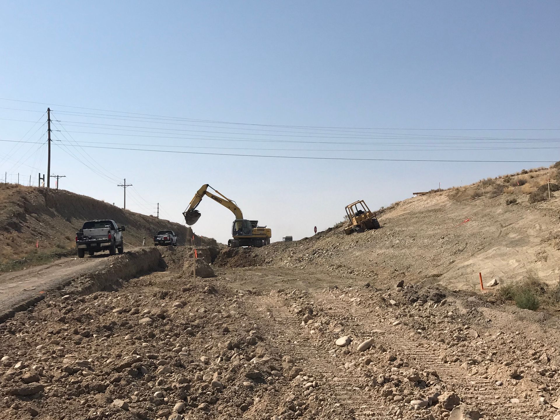Construction site on dirt road; excavator and small dozer moving earth. Car driving nearby under clear sky.