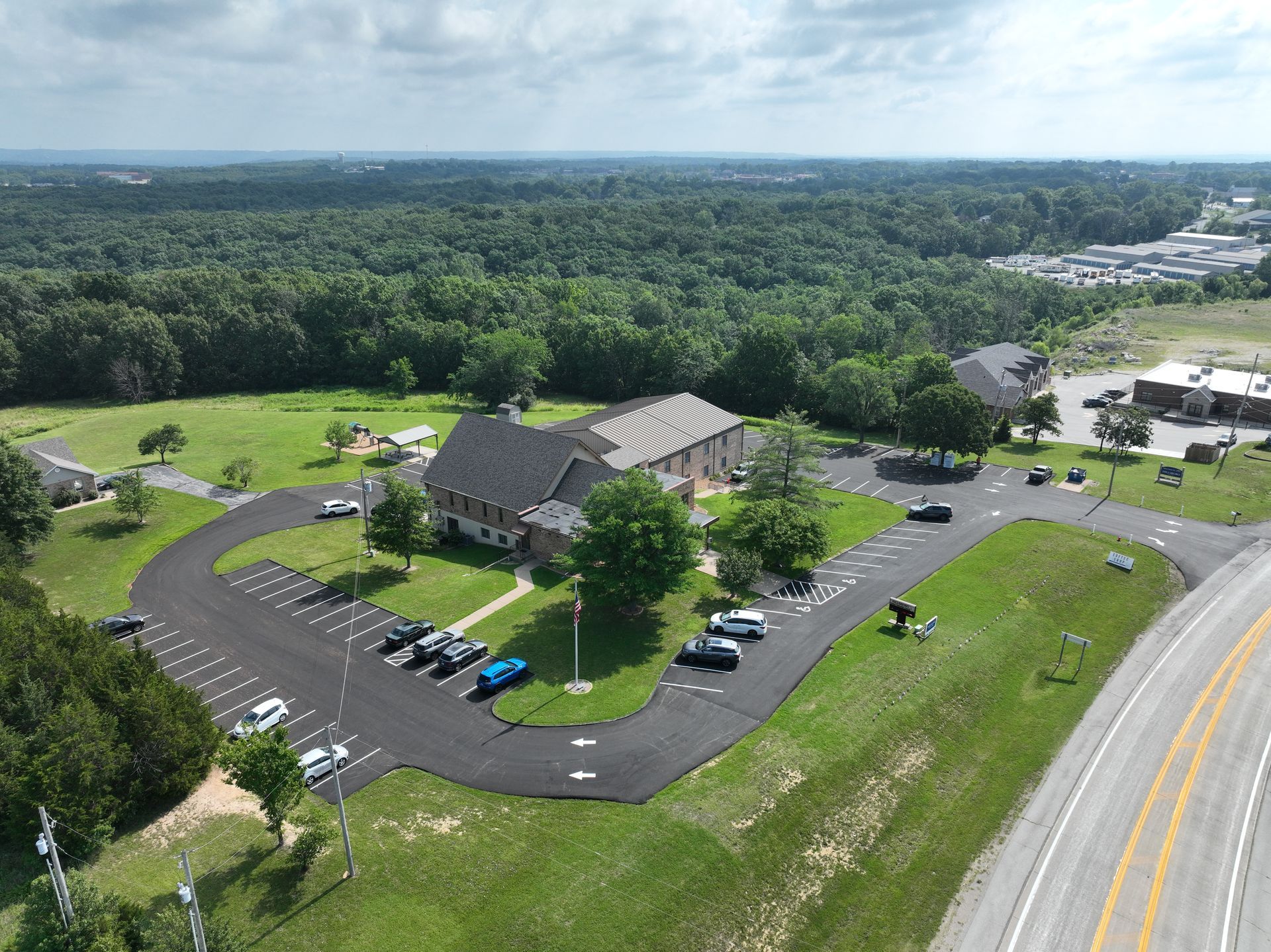 Aerial view of a building with parking lot, trees, and a highway beside a wooded area.