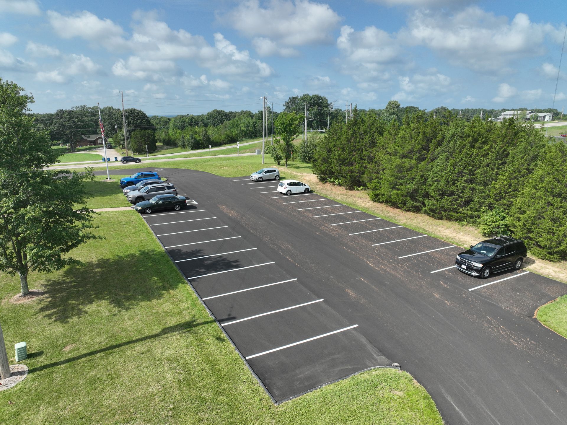Parking lot with several cars beside a grassy field and trees under a partly cloudy sky
