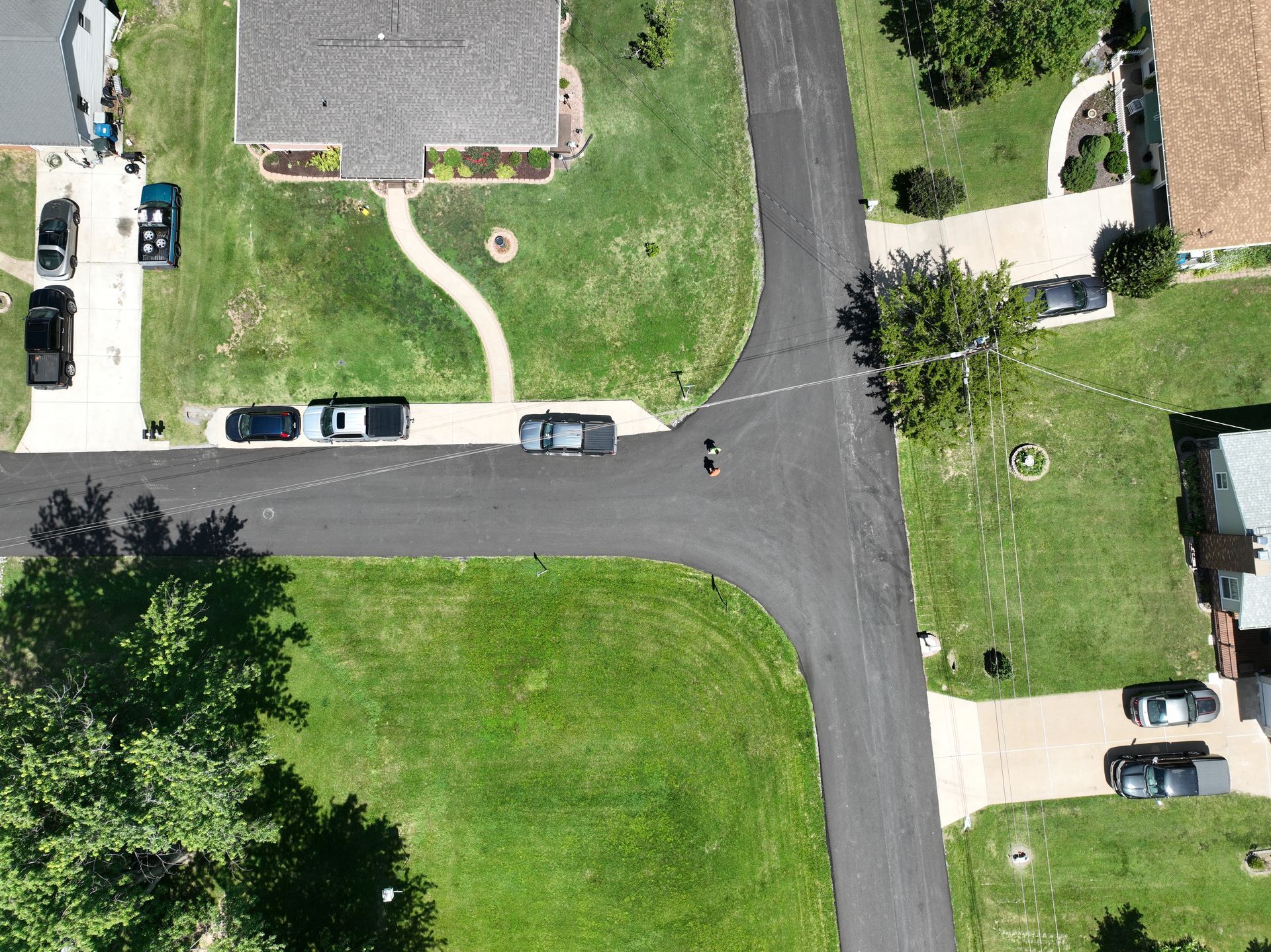 Aerial view of a suburban street intersection with houses, lawns, sidewalks, and parked cars.