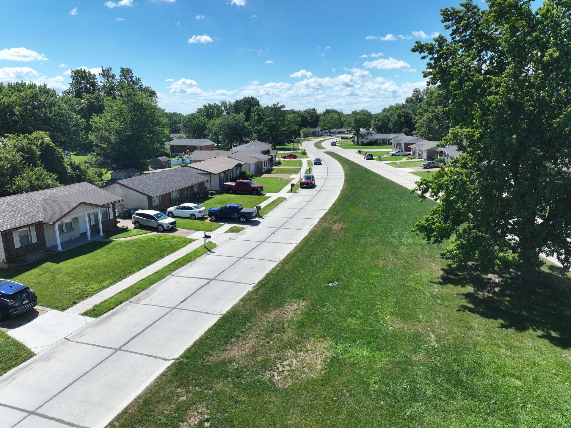 Aerial view of a suburban street with houses, parked cars, and green lawns under a blue sky.