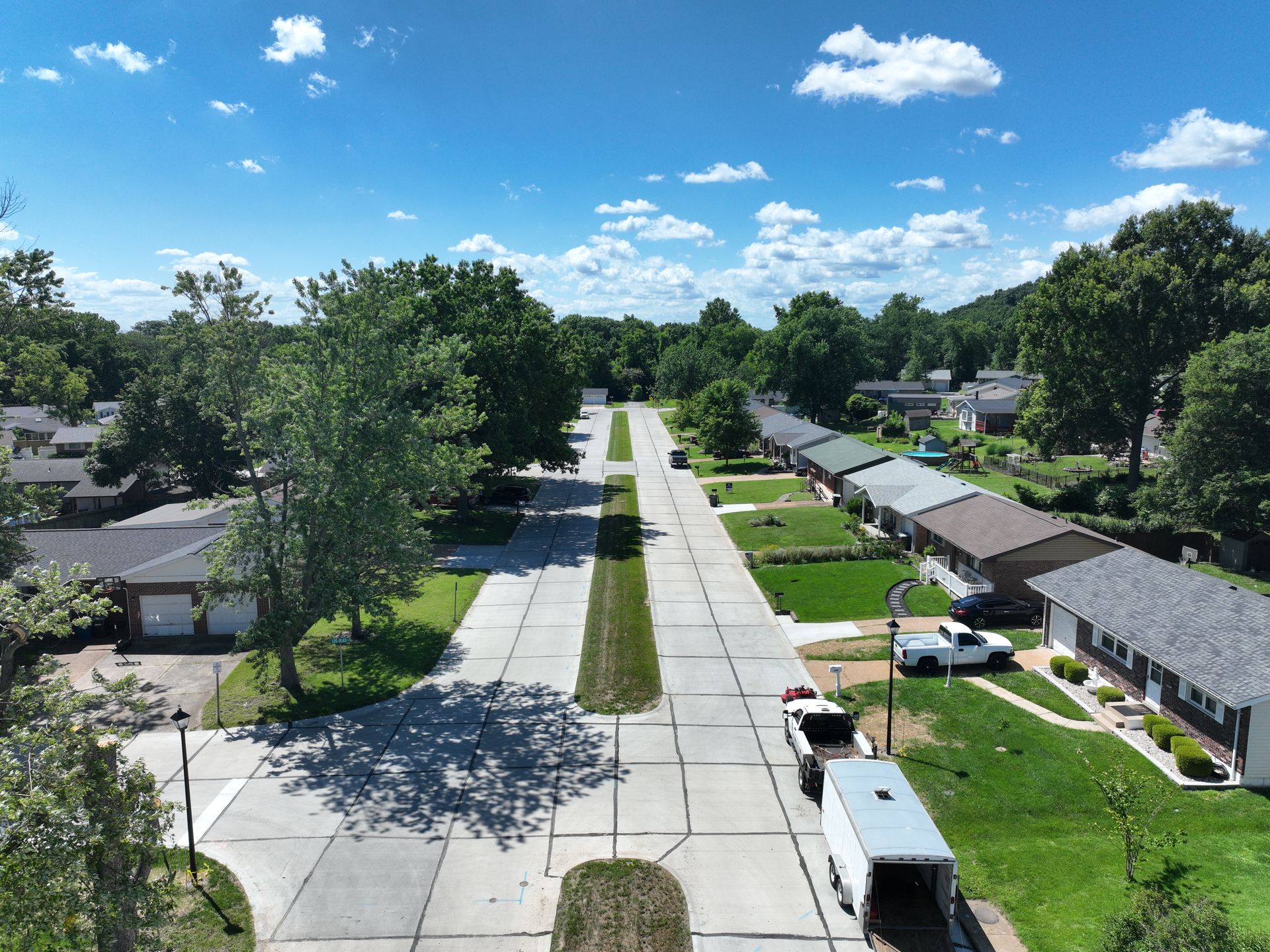 Aerial view of a quiet suburban street with houses, trees, parked cars, and blue sky.
