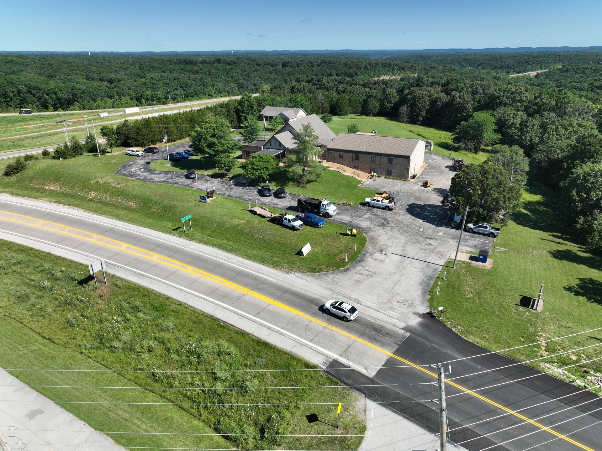 Aerial view of a rural building complex beside a road, surrounded by trees and parking area