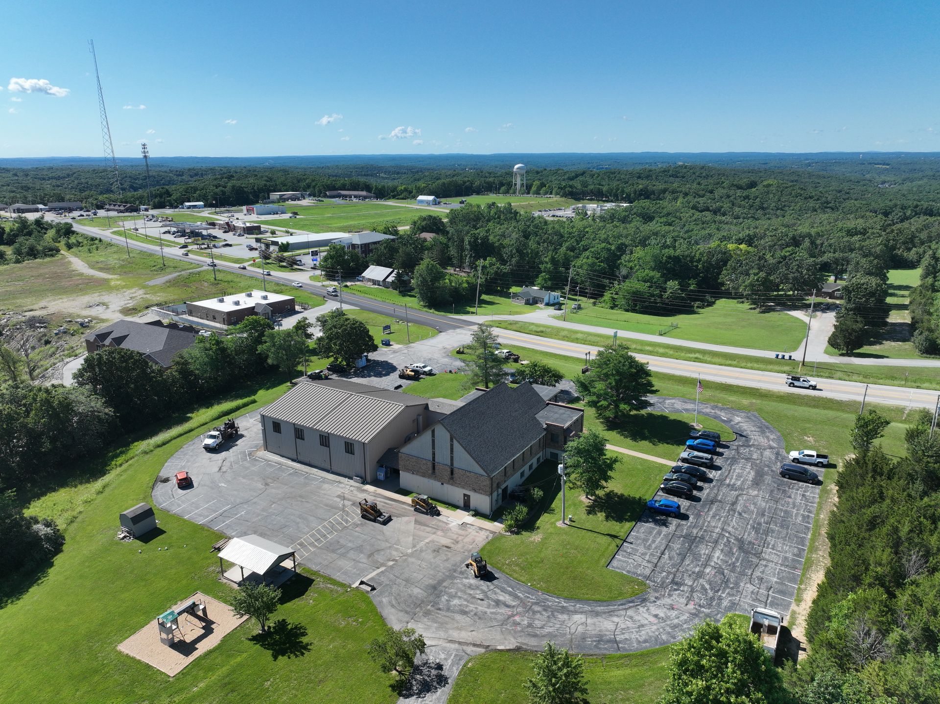 Aerial view of a brown-roofed building, parking lot, and surrounding green fields by a road.