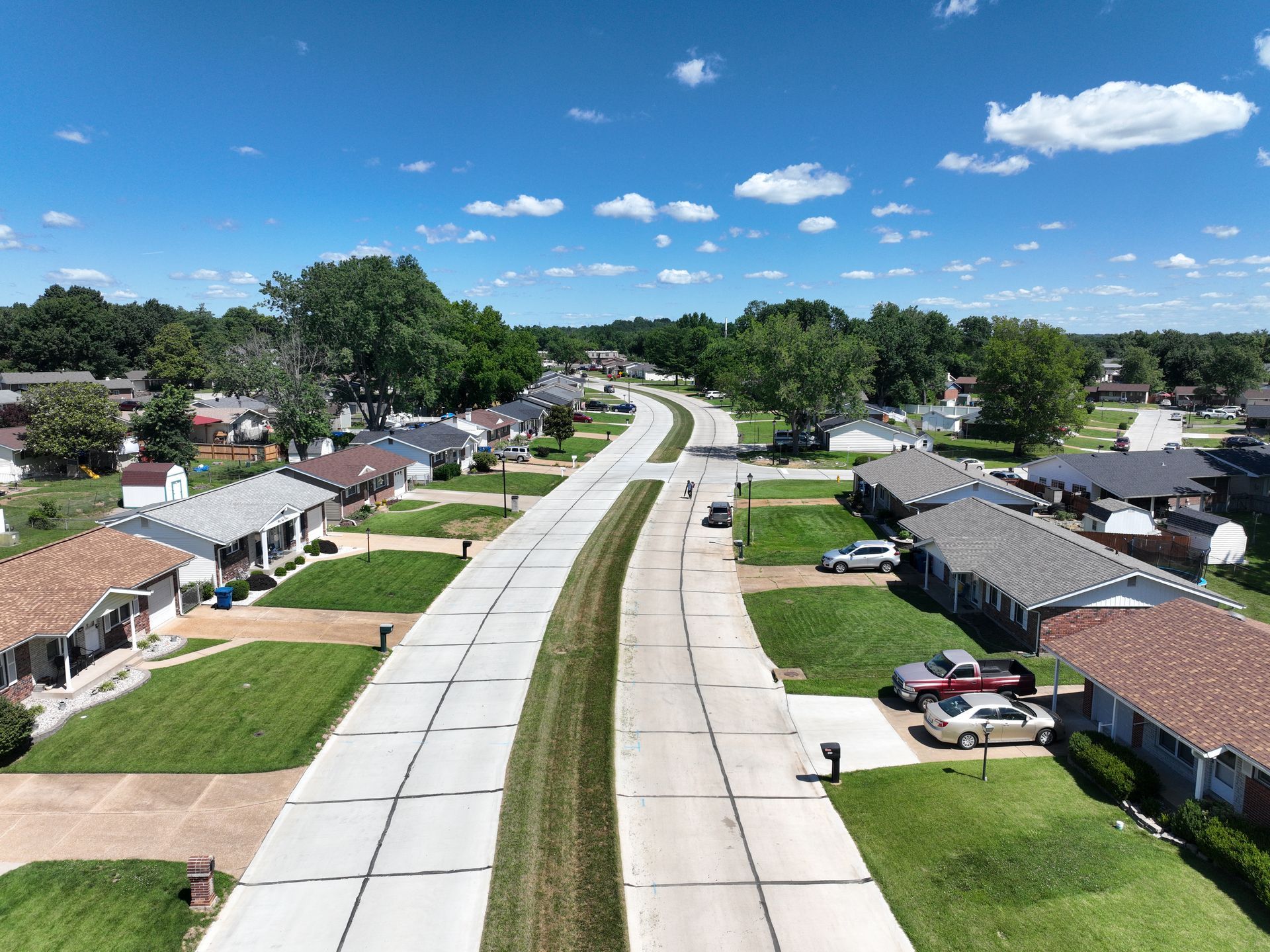 Aerial view of a suburban street with houses, lawns, and a blue sky with scattered clouds.