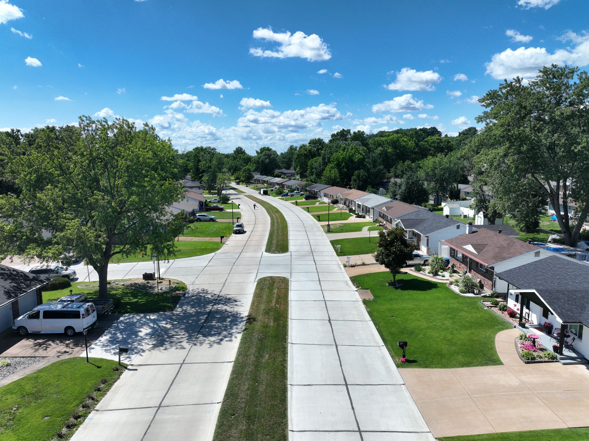 Aerial view of a suburban street with houses, trees, and a bright blue sky with scattered clouds