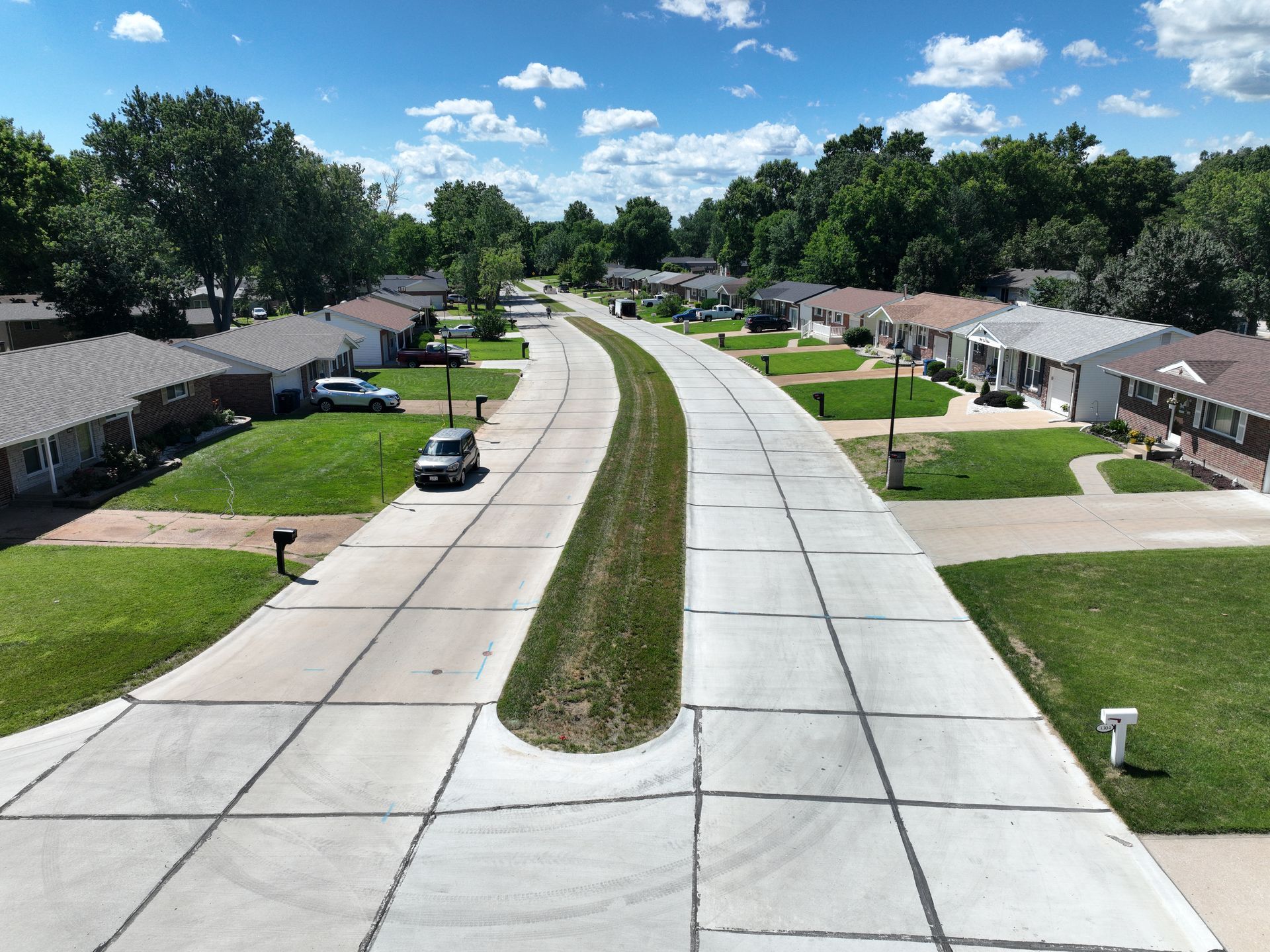 Suburban street with homes on both sides, curving concrete road, grassy median, and trees under a blue sky