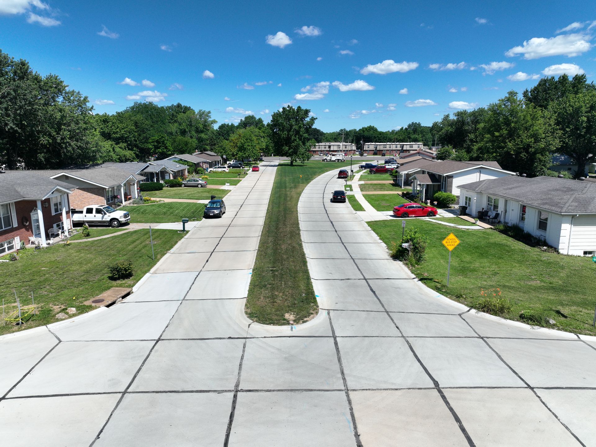 Suburban neighborhood street with sidewalks, green lawns, and houses under a bright blue sky