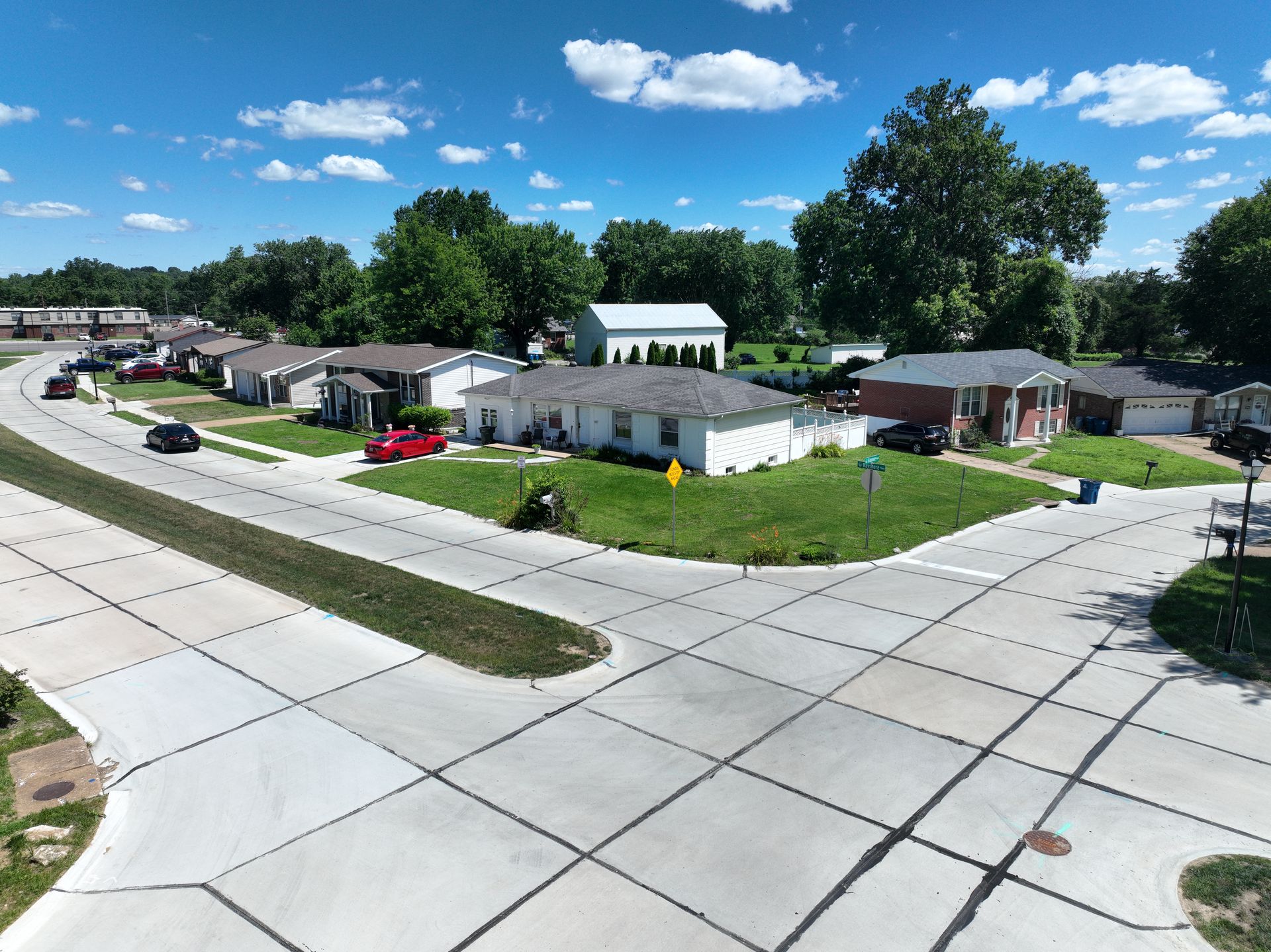 Aerial view of a suburban neighborhood with houses, streets, green lawns, and a blue sky with clouds.