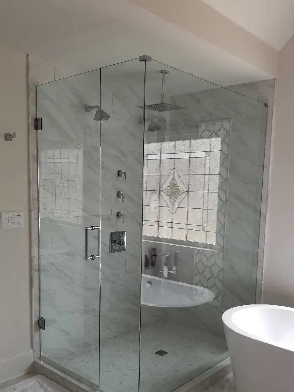 Glass shower enclosure in a bright bathroom with tiled walls and a freestanding tub nearby