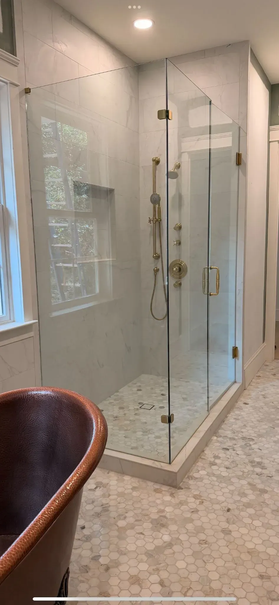 Glass shower enclosure in a bright bathroom with beige tile and a wicker chair in the foreground