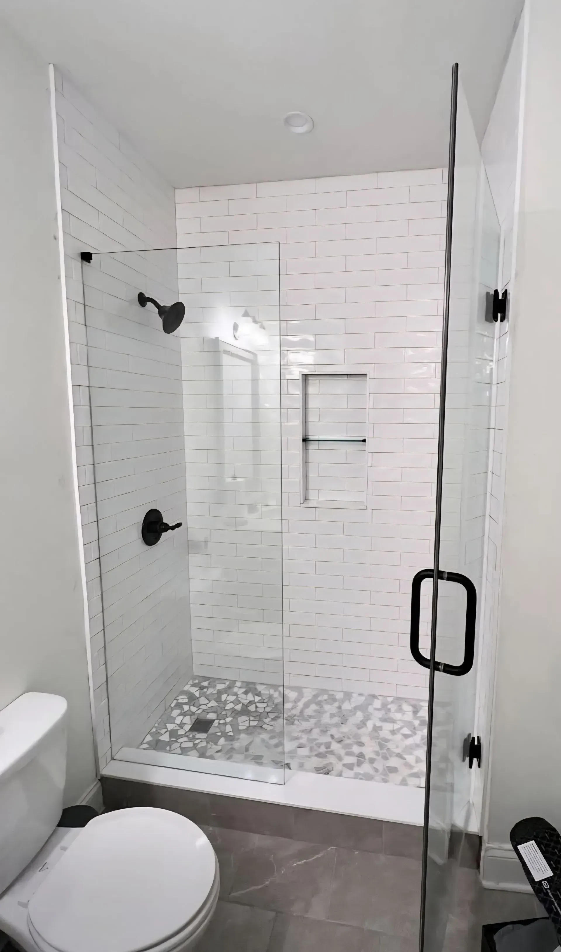 Small white-tiled bathroom with a glass-enclosed shower, toilet, and black fixtures.