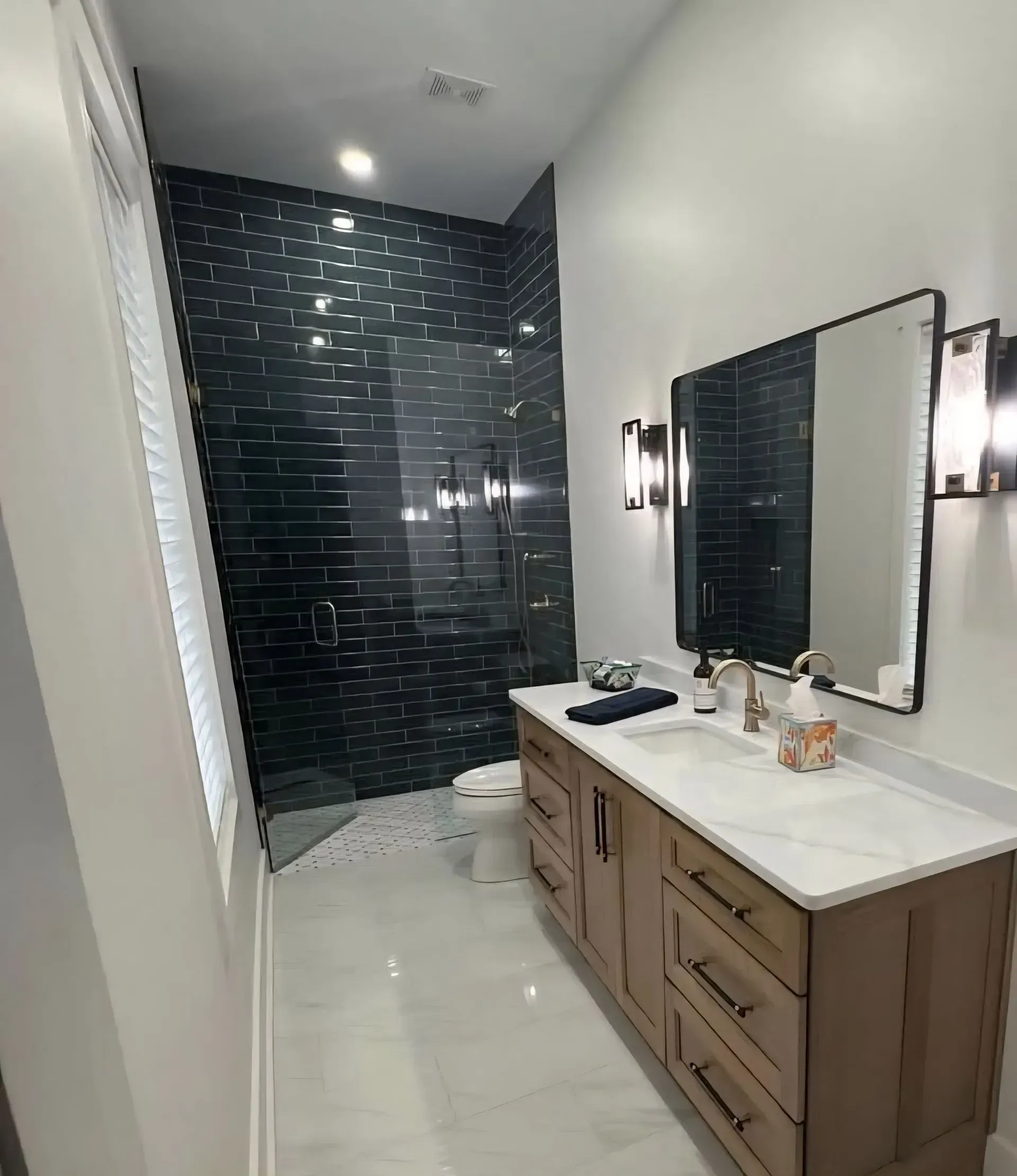 Modern bathroom with dark tiled shower, white countertop, wooden vanity, and mirror sconces.