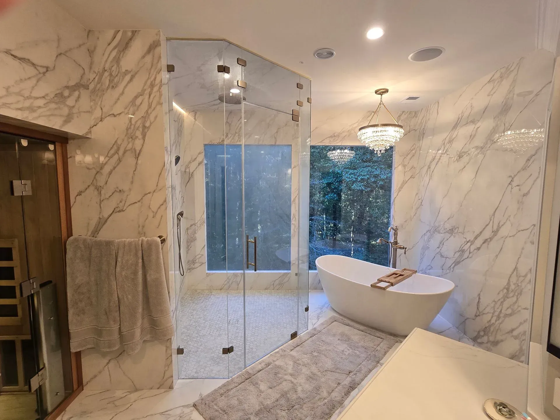 Modern bathroom with marble walls, glass shower, freestanding tub, and large window overlooking trees