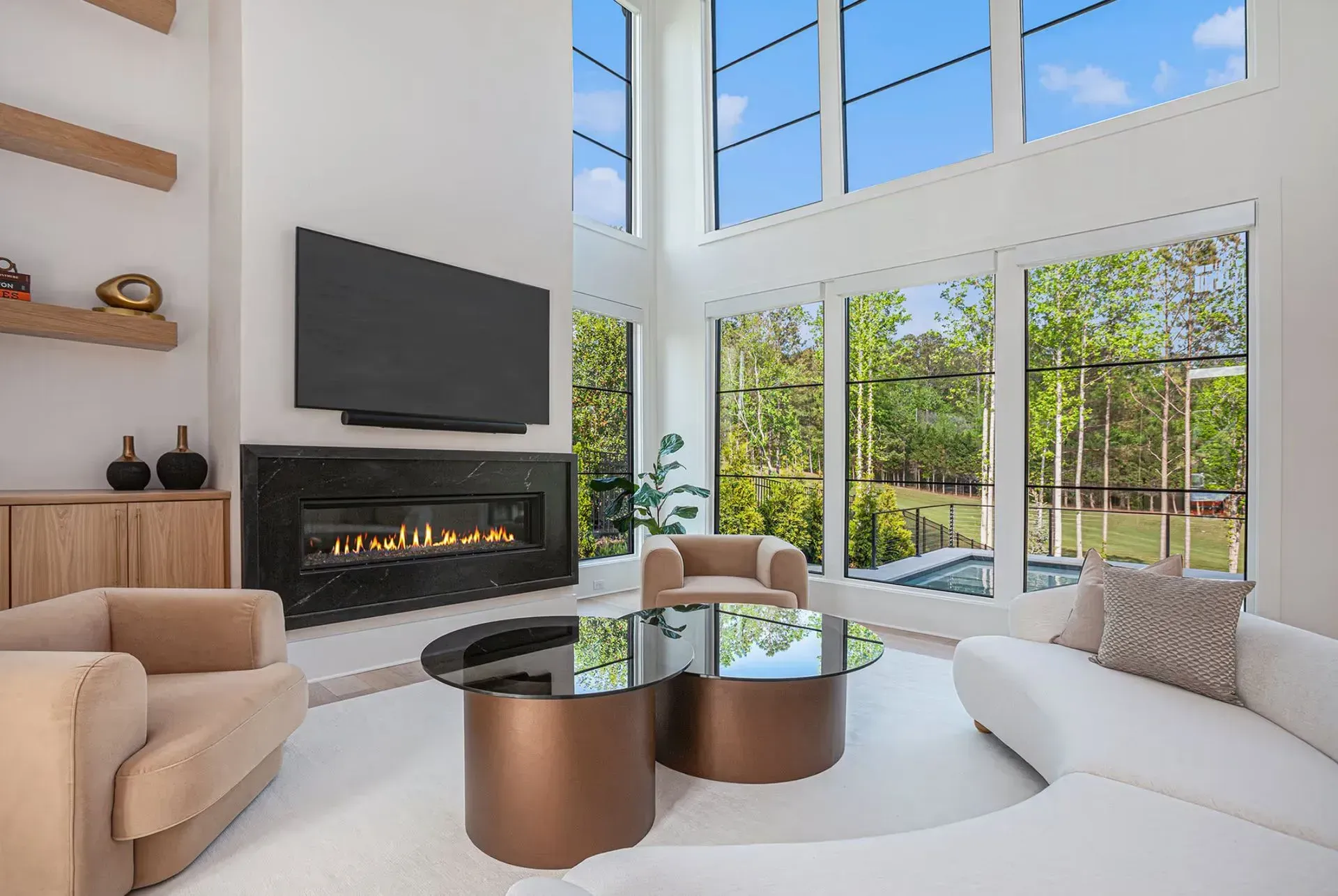 Modern living room with white seating, glass coffee table, fireplace, TV, and tall windows overlooking greenery