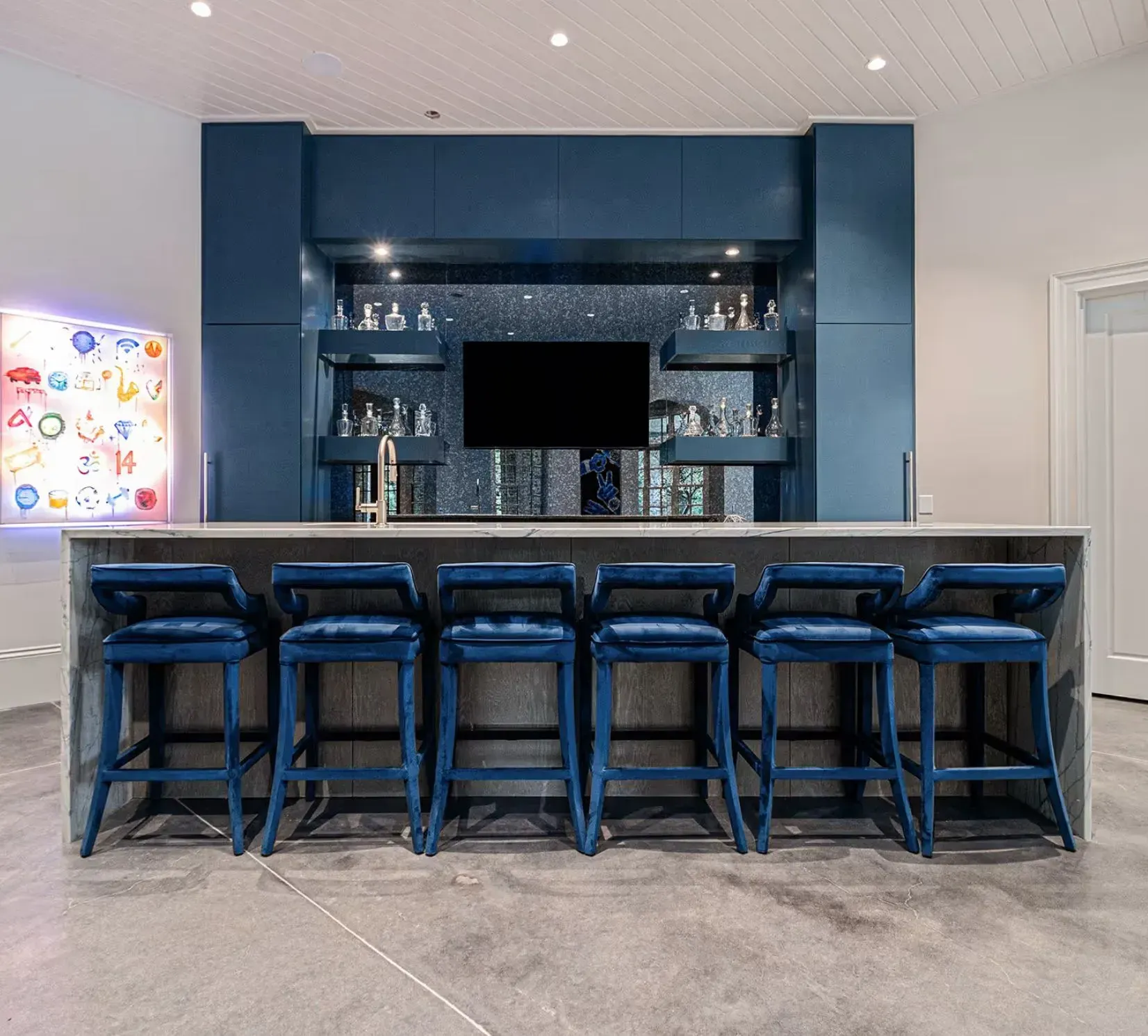 Modern blue home bar with five stools, mirrored shelves, and a wall-mounted TV against a light interior.
