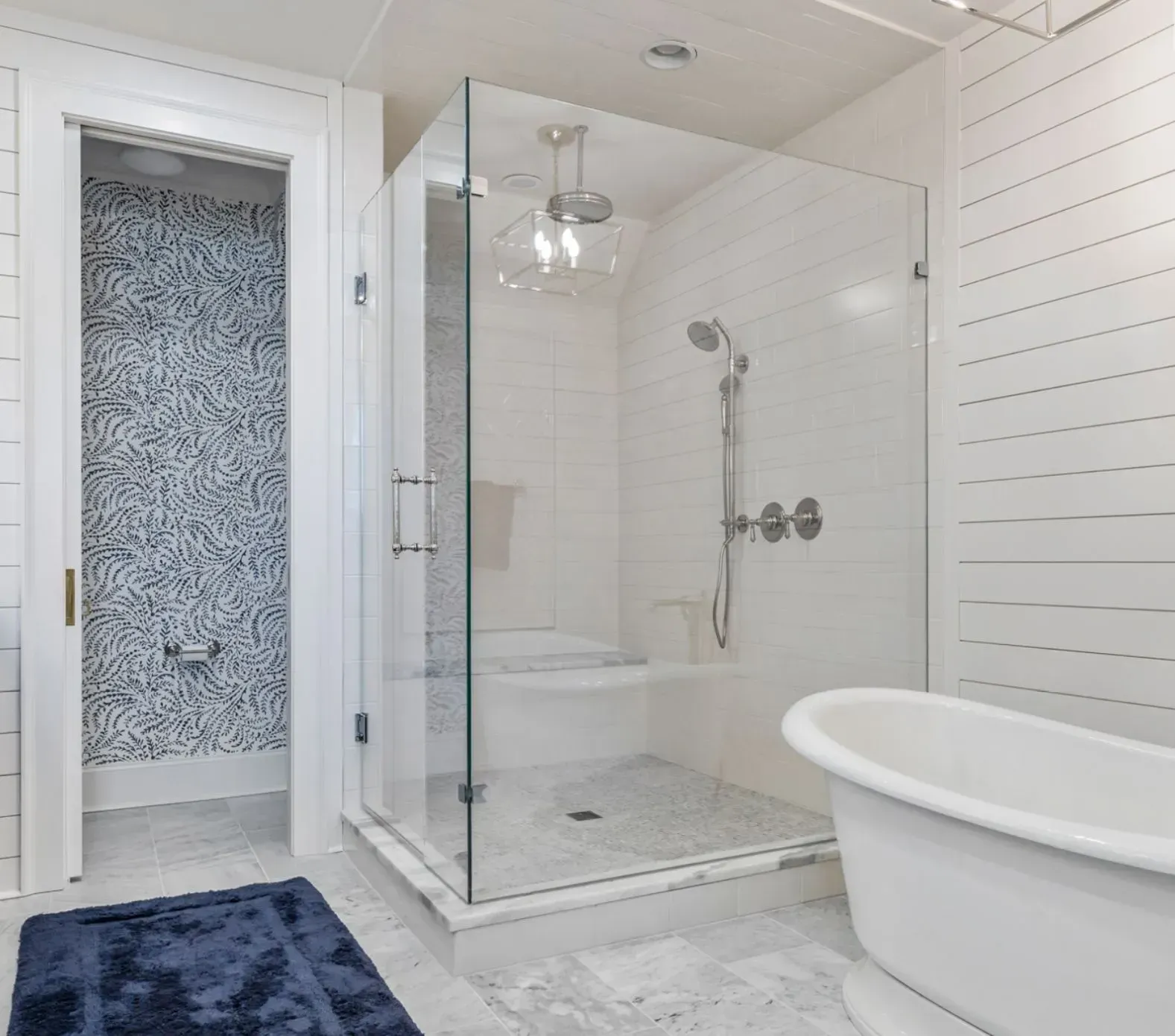 Bright white bathroom with glass shower, freestanding tub, frosted door, and dark blue rug
