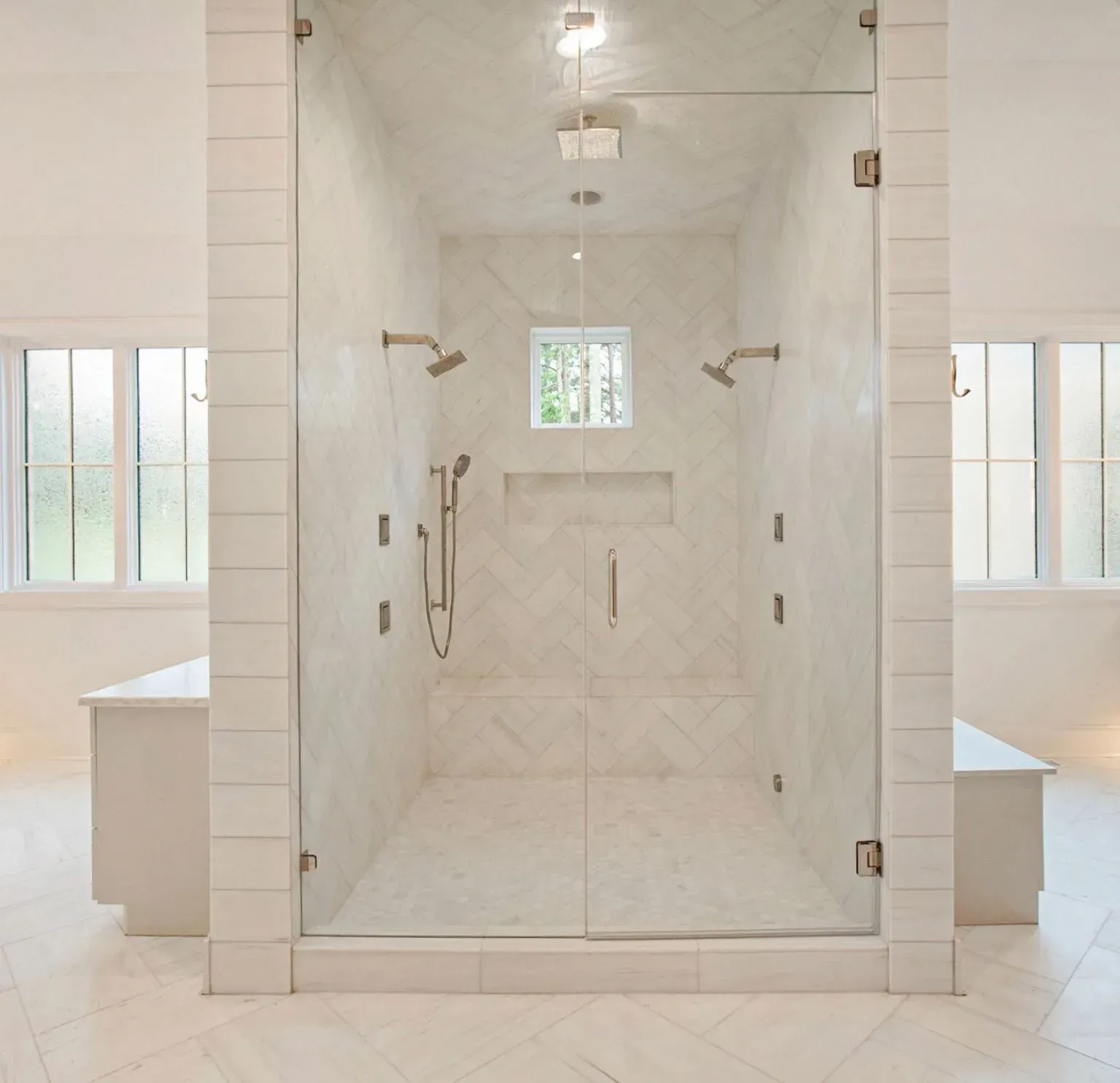 White marble shower with glass door, built-in bench, grab bars, and small window in a bright bathroom.