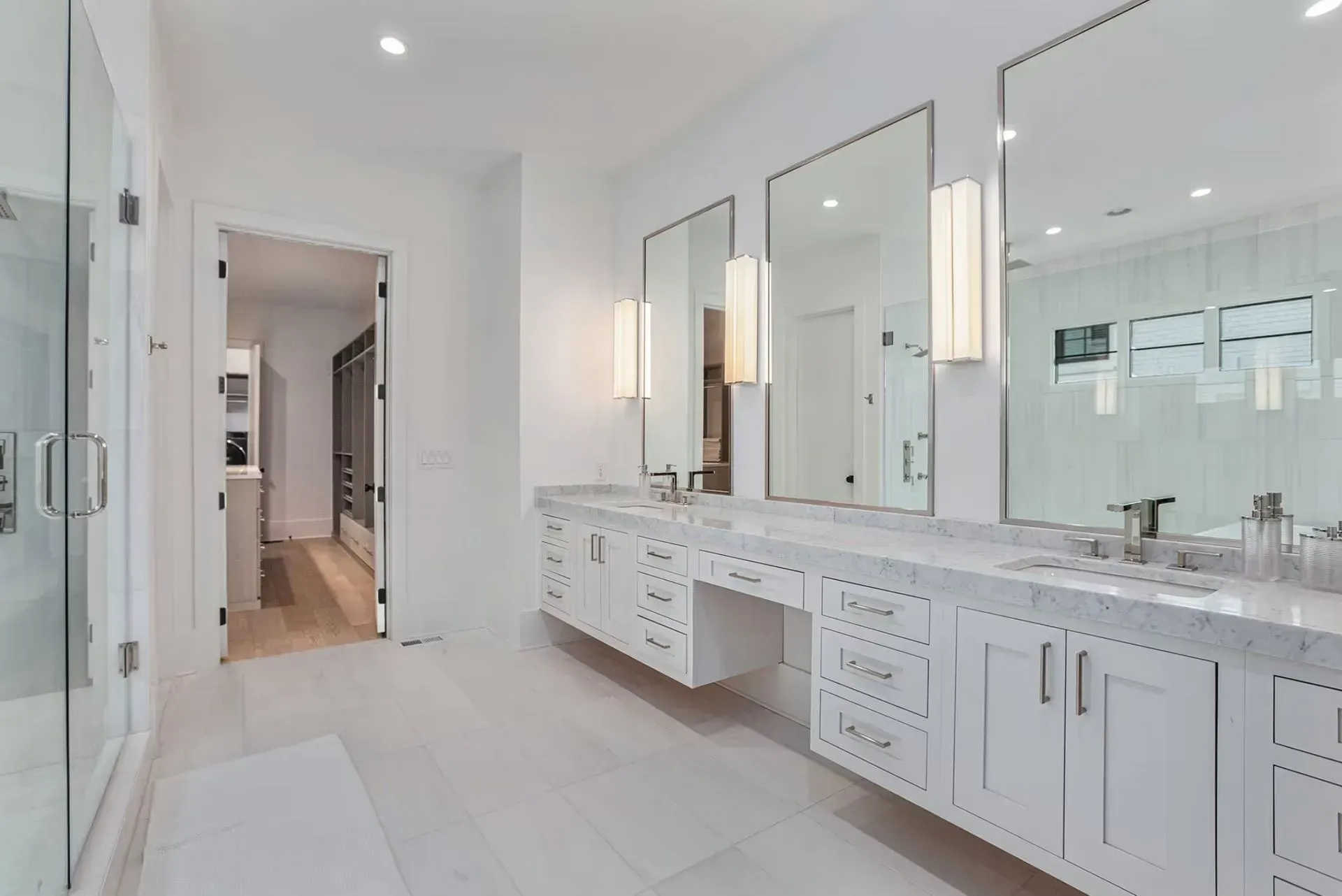 Bright white bathroom with double vanity, large mirrors, and a doorway leading to a hallway.