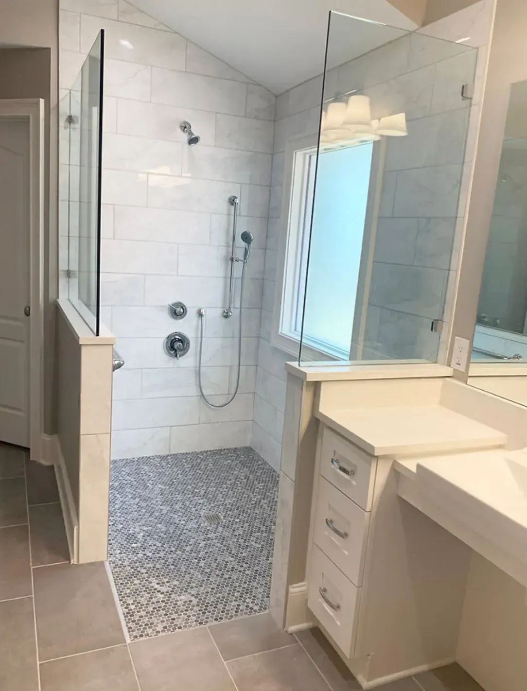 Modern bathroom with white-tiled walk-in shower, glass enclosure, gray pebble floor, and vanity sink.