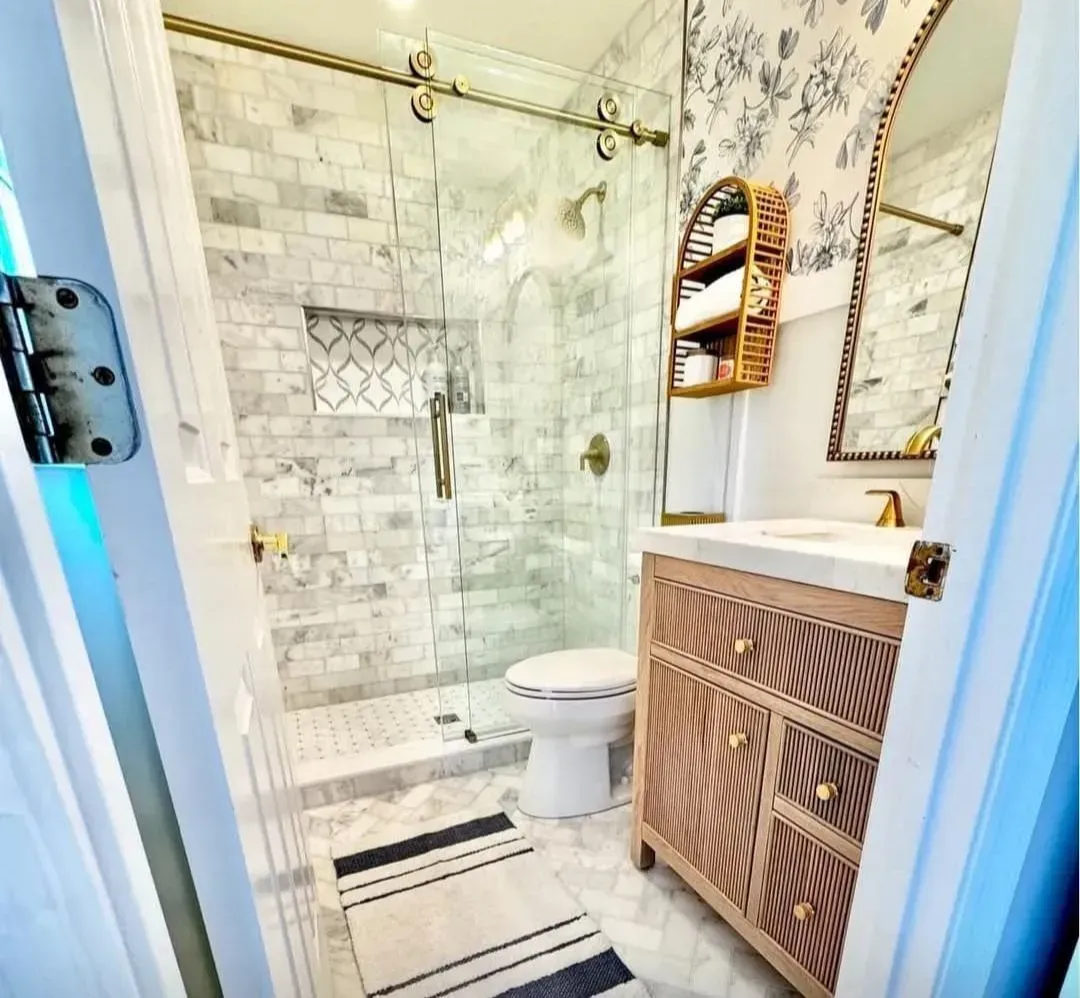 Bright bathroom with glass shower, white tile, striped rug, and wooden vanity with round mirror.