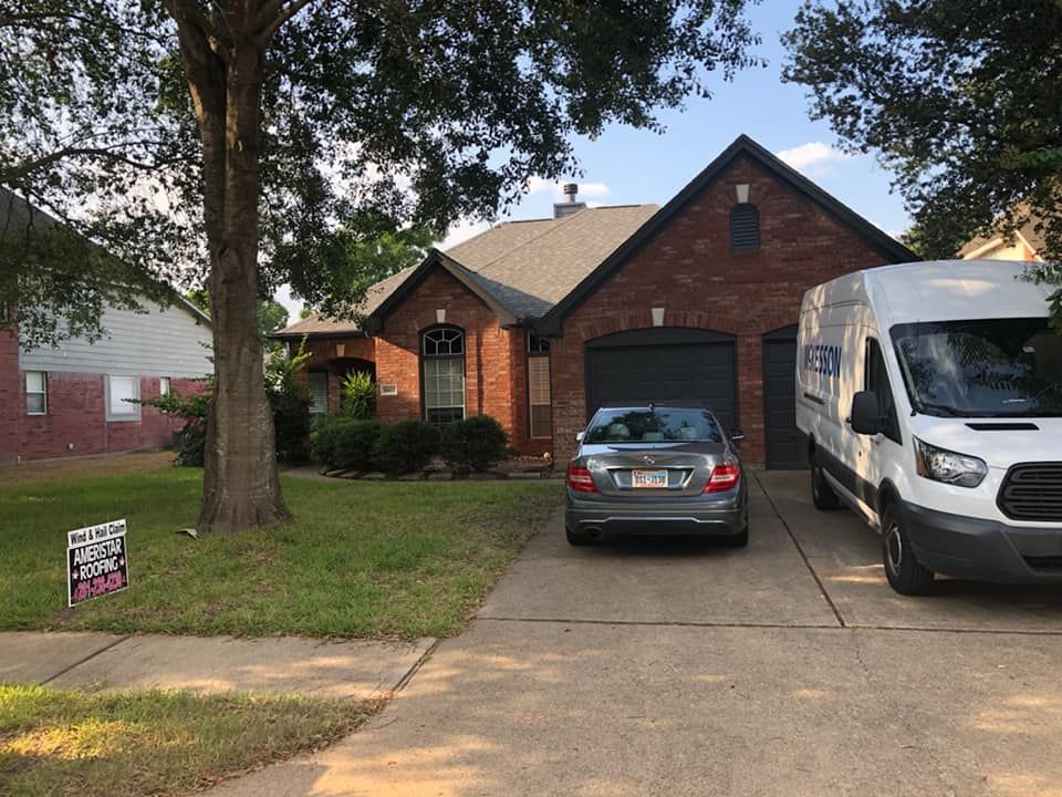 Brick house with gray garage door, a car, and a white van parked in the driveway. Green lawn and trees in front.