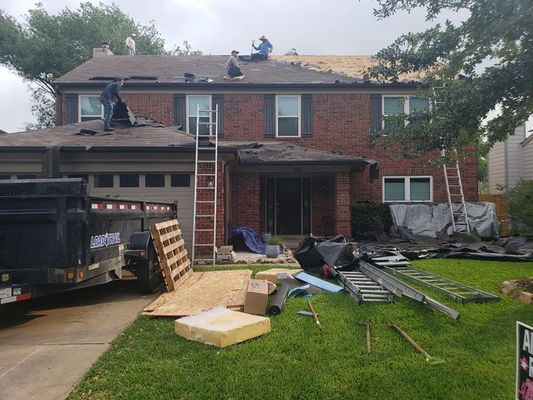 Roofers working on a two-story brick house, removing old shingles and laying new ones. Ladders, tools, and materials are on the lawn.