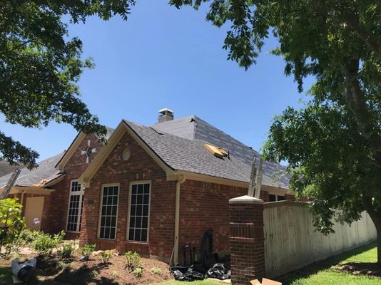 A brick house with a dark gray roof under a blue sky, framed by green trees. A wooden fence runs along the side.
