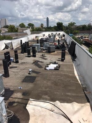 Flat commercial roof with HVAC equipment and tall cylindrical black objects, under a cloudy sky. Buildings in the distance.