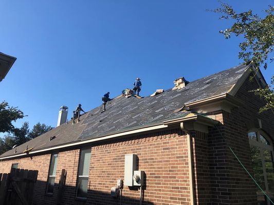 Workers replacing shingles on a brick house roof under a clear blue sky. Several people are visible on the roof, and the old shingles are partially removed.
