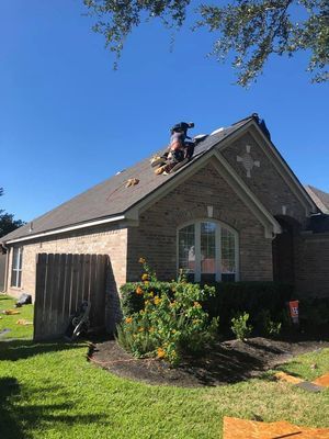 Workers replacing shingles on the roof of a brick home on a sunny day. Green grass and bushes surround the house.