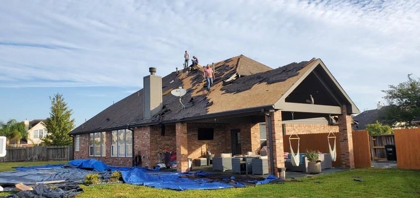 Workers removing roofing shingles from a brick house with a partially covered deck. Blue tarps cover the yard.