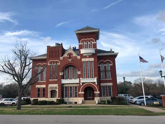 Red brick courthouse with a central tower and arched windows under a cloudy sky. Two American flags fly outside.
