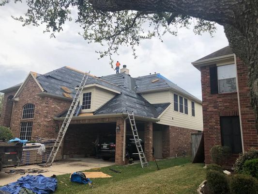 Roofers work on a house with brick and siding. Ladders lean against the roof, a car is parked in the garage.