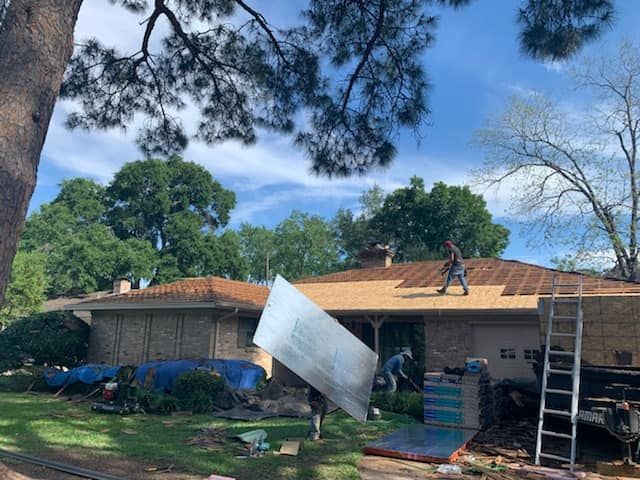 Workers installing roofing shingles on a one-story house with a partially exposed roof. Sunny day with green trees.
