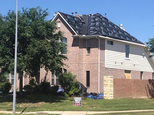 House with a partially covered roof. Brick exterior, white siding, and tarp visible on the damaged roof under a blue sky.