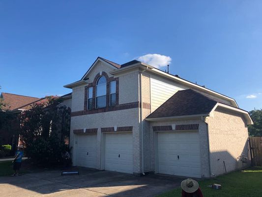 Two-story house with beige stucco and brick, three-car garage, and dark brown roof against a blue sky.