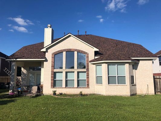 Back view of a light-colored brick house with a brown roof and large windows, under a clear blue sky. Green grass in the foreground.