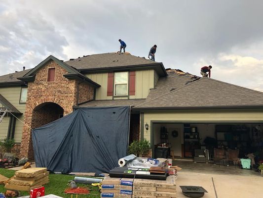 Three roofers working on a two-story house with visible damage and materials on the lawn; blue tarp covers the garage entrance.
