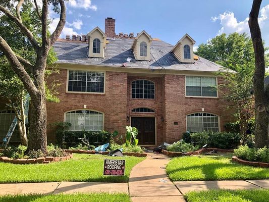 A two-story brick house with roof damage, debris on the lawn, and a for-sale sign.