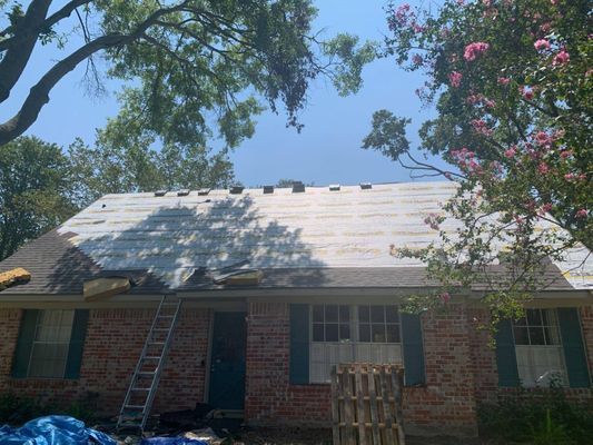 Roof of a brick house undergoing repair with shingles removed. A ladder leans against the house, blue tarps are below.