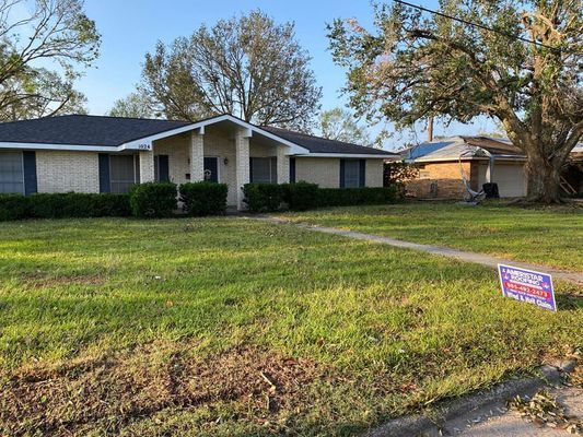A single-story brick house with a green lawn and a political sign in front. A second building is visible to the right.
