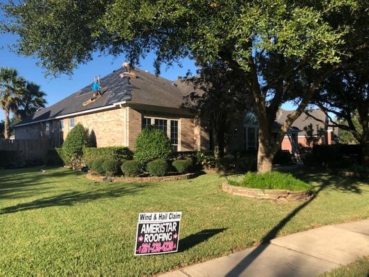 House with roofing work in progress, including a worker on the roof, on a sunny day with a sign in the front yard.