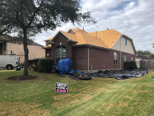 House with a partially completed roof renovation, covered in orange roofing and tarps, set against a cloudy sky and green lawn.