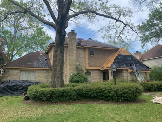 A house with a partial roof replacement, a worker on a ladder, a tree, and a brick facade.