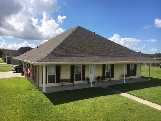 A light yellow house with a brown roof and a long porch, on a grassy lawn under a blue sky with clouds.