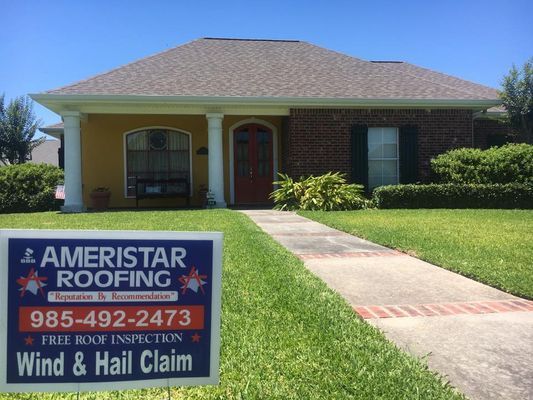 A sign for Ameristar Roofing in front of a house with a brick and yellow exterior and a green lawn.