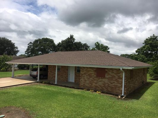 A one-story brick house with a brown roof and carport sits on a green lawn under a cloudy sky.