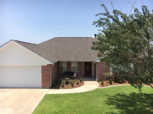 A brick house with a brown roof, white garage door, and red front door. Green grass and a tree are in the foreground.